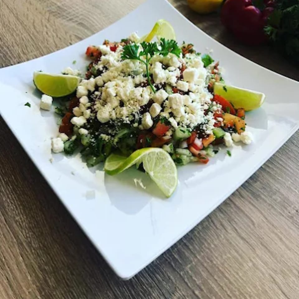 Turkish salad with diced tomatoes, cucumbers, feta cheese, parsley, and lime wedges on a white plate.